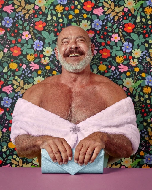 Bald adult male smiles in front of a floral wallpaper background wearing a pink wrap and holding a blue clutch purse.